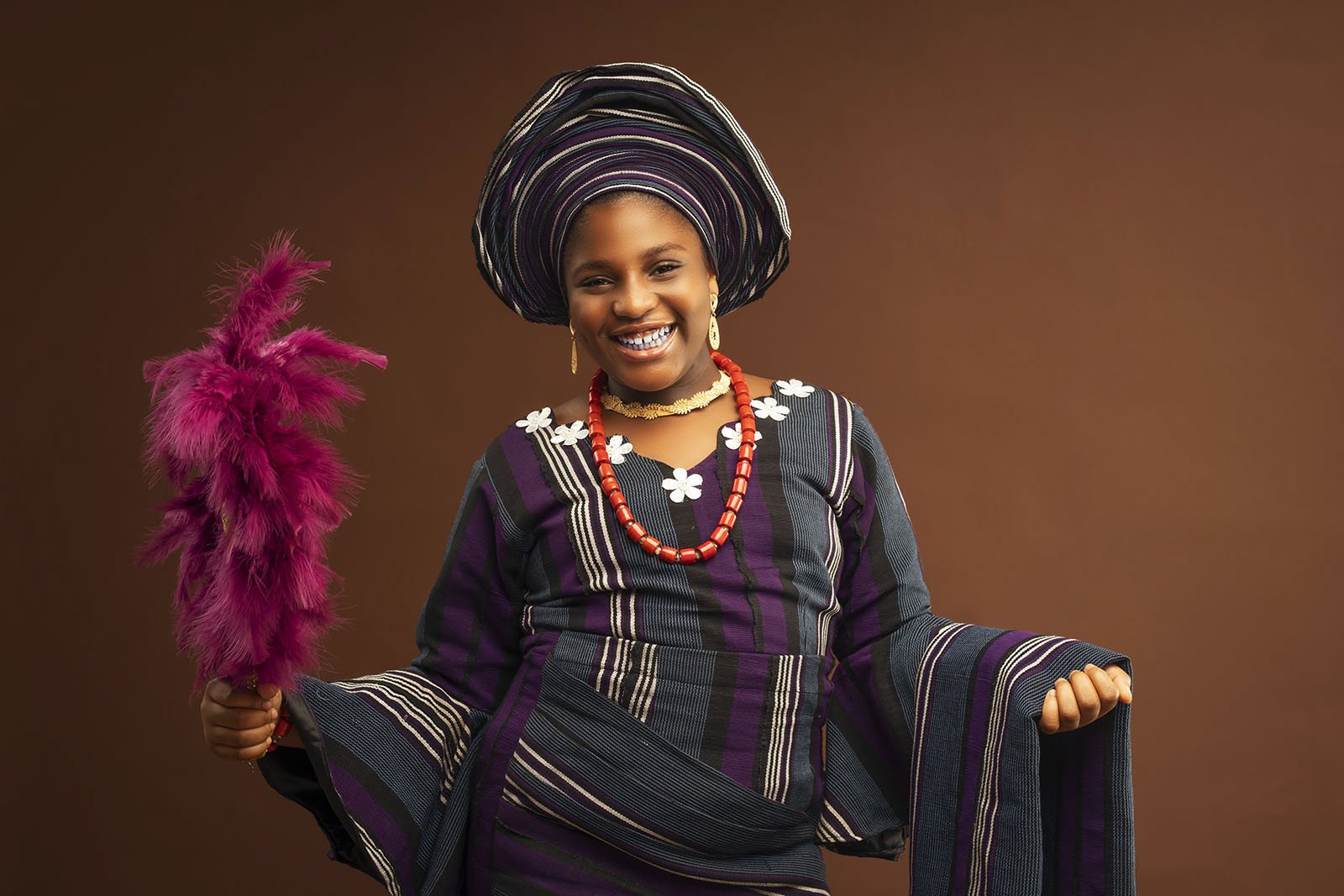 Smiling woman in traditional aso-oke attire holding a pink feather fan